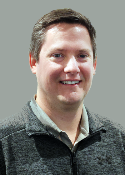 Corporate portrait of a smiling man in a dark zip-up sweater with a light collared shirt underneath.