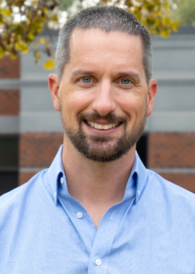 Professional outdoor headshot of a smiling man wearing a light blue button-down shirt in front of an office building.