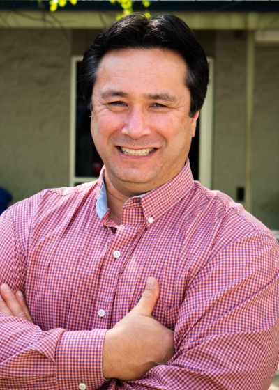 Professional outdoor portrait of a smiling man wearing a red checkered button-down shirt with arms crossed in front of an office building.
